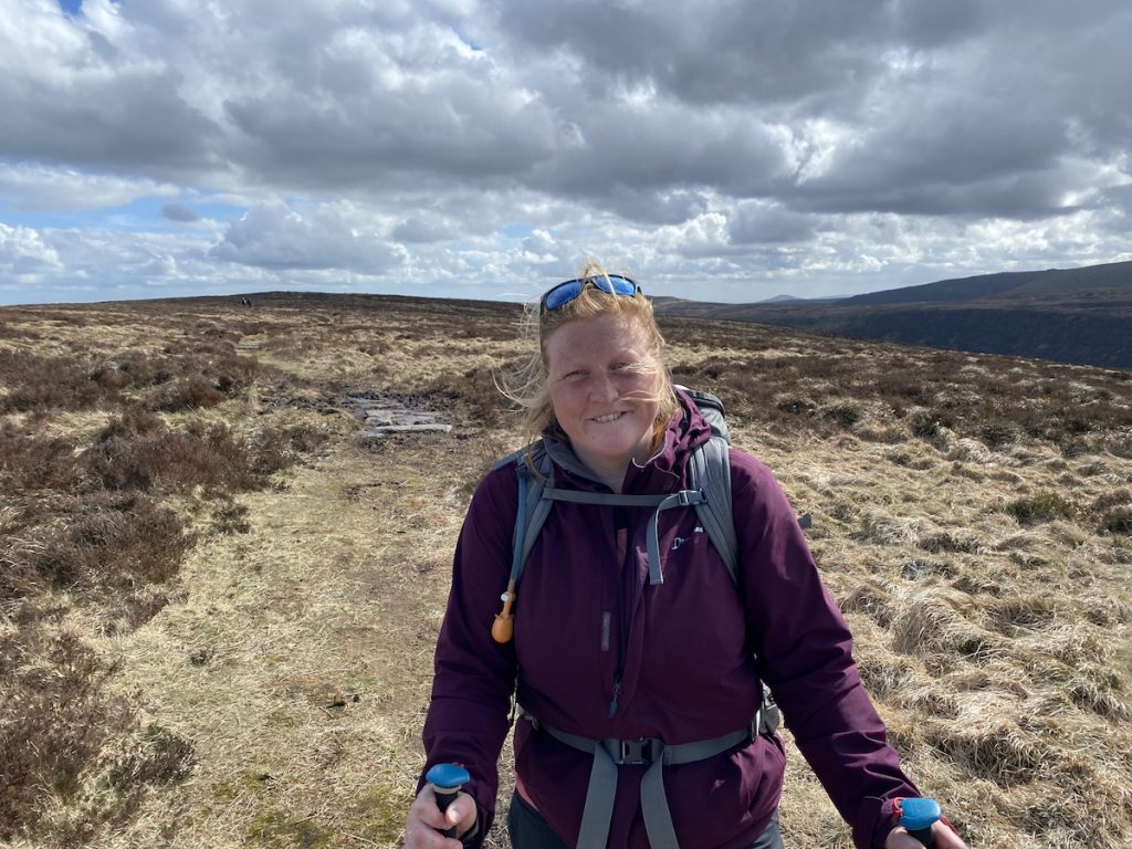 A winswept lady with ginger hair wearing waterproofs and a rucksack stood on a hill side