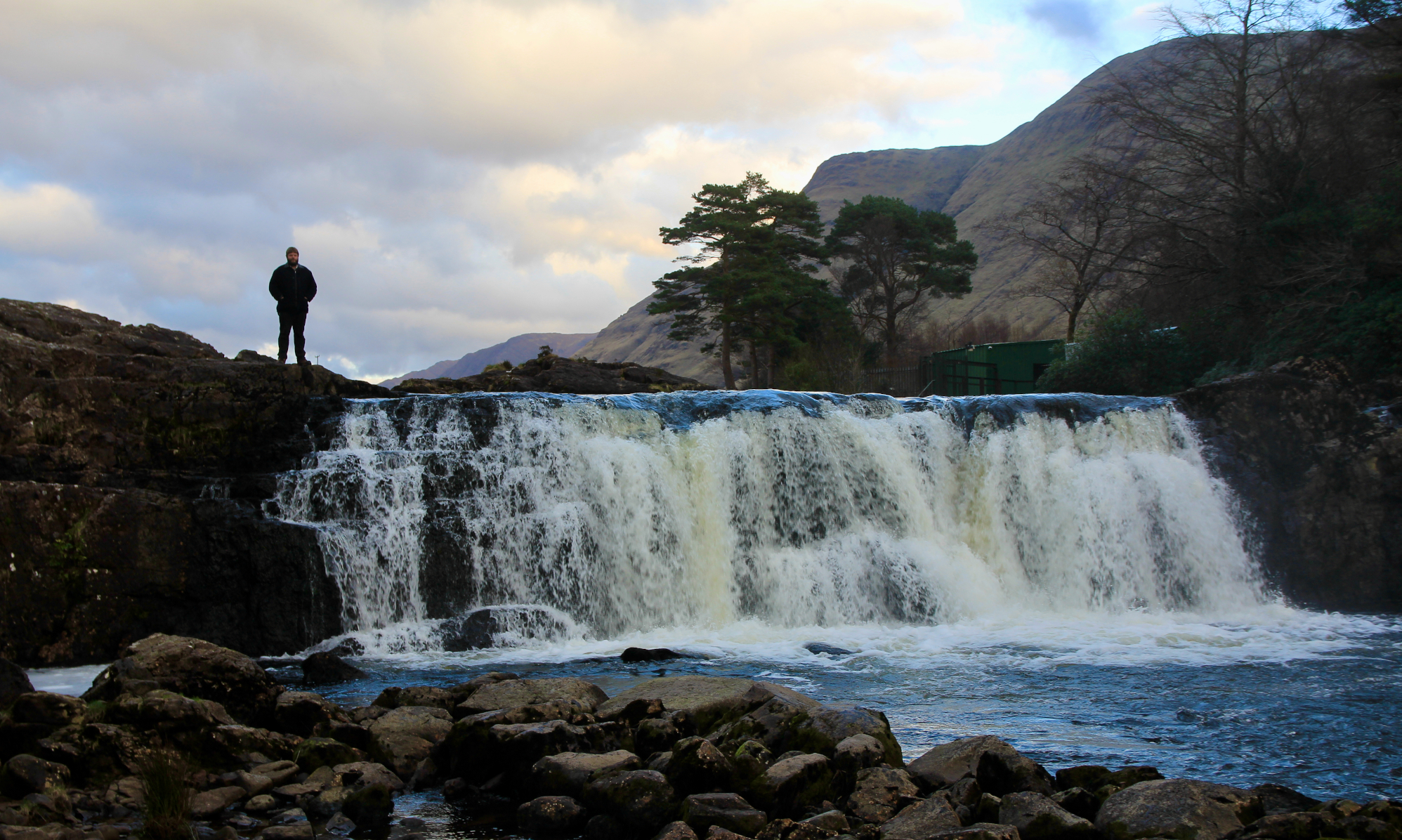 Outdoor Instructor stood on the top of a wide waterfall with mountains in the background practicing navigation in his adventures and answering faq