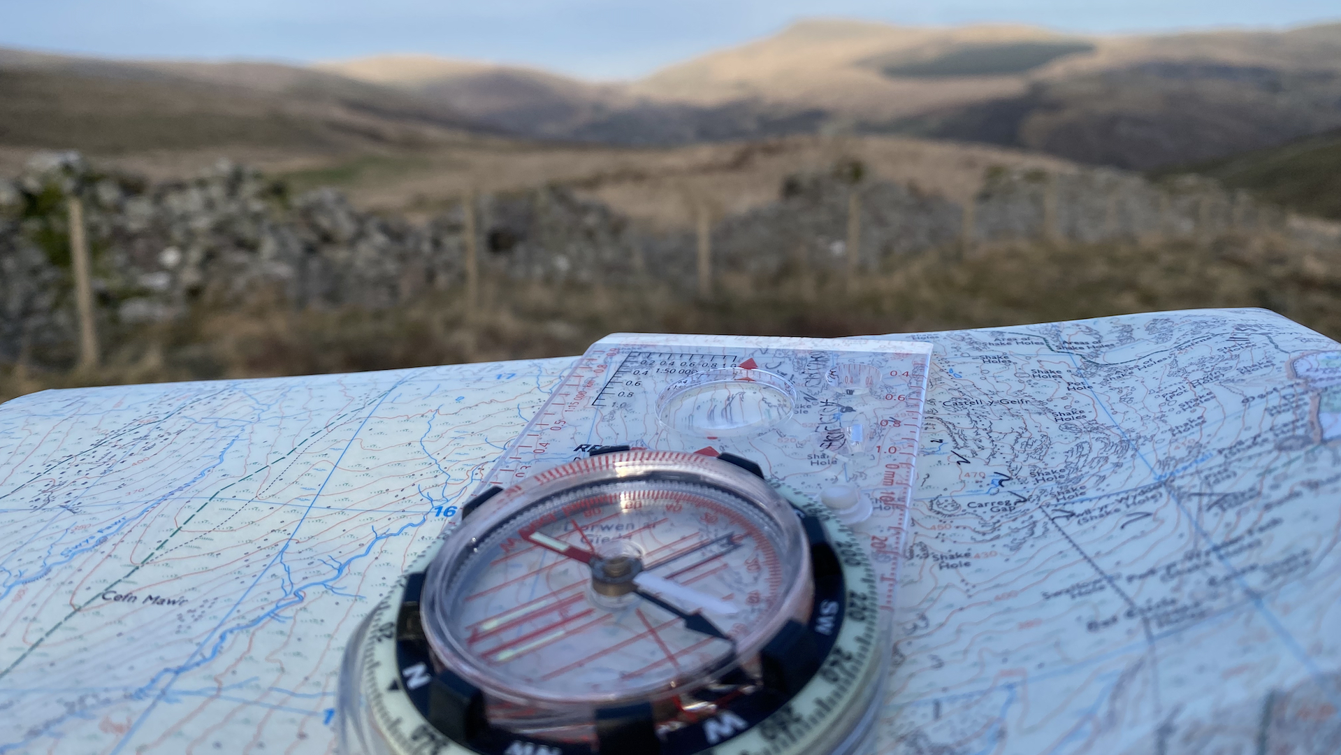 An image of an ordnace survey map and compass in the foreground, with a drystone wall and hills in the distance. A map and compass that would be used on the Introduction to Navigation course.